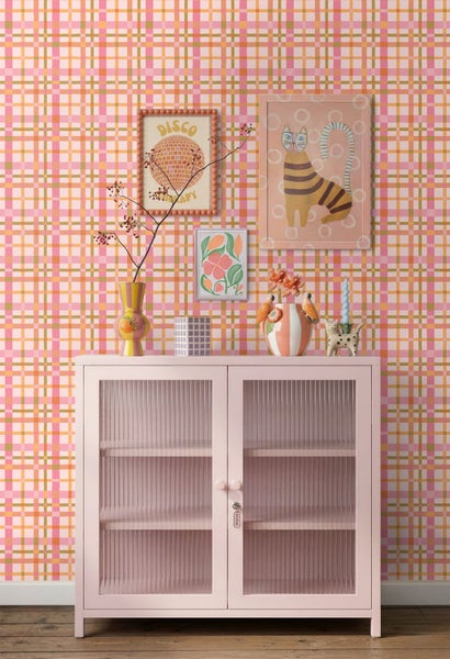 Vliestapete mit Karomuster in Rosa, Orange und Grün hinter einer rosa Kommode mit Glasfront und Dekoration.