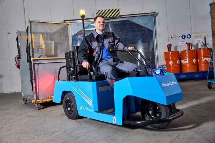 Mitarbeiter fährt einen blauen HanseLifter Elektro-Schlepper mit Lithium-Ionen-Batterie im Lager und zieht einen Metallcontainer.
