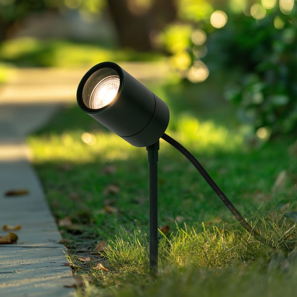 Schwarzer LED-Gartenstrahler mit Erdspieß in einer Grünanlage neben einem Gehweg, eingeschaltet mit warmem Licht.