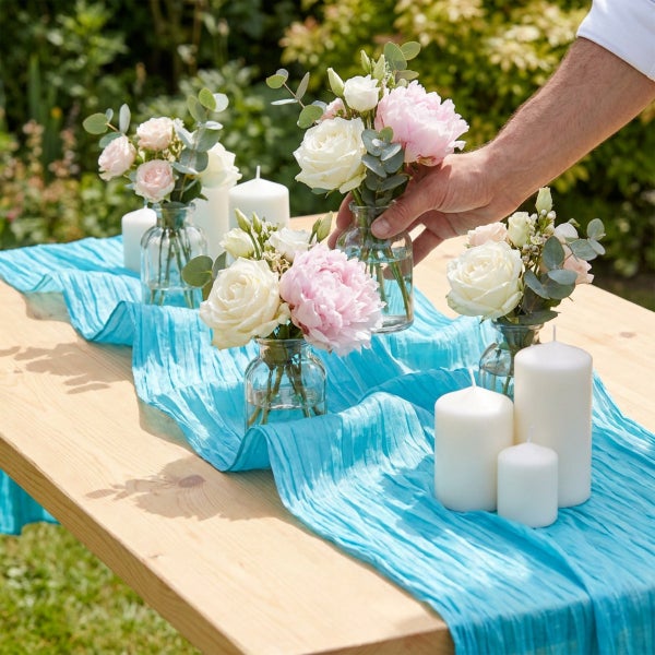Holztisch dekoriert mit Glasvasen voller Blumensträuße, Stumpenkerzen und einem strukturierten Tischläufer im Freien.
