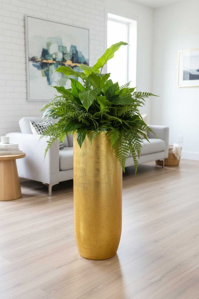 Hoher zylindrischer goldfarbener Pflanzkübel mit grünen Pflanzen in einem hellen, modernen Wohnzimmer mit Sofa und Holzboden.