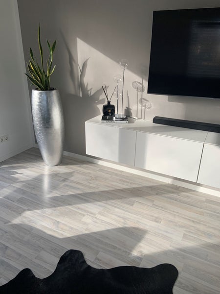 Helles Wohnzimmer mit Bodenbelag in Holzoptik, weißem schwebendem Lowboard, großer silberner Bodenvase mit Bogenhanf und schwarzem Teppich.
