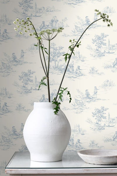 Vliestapete mit Toile-de-Jouy-Muster in Blau und Weiß, davor eine weiße Vase mit dekorativen Pflanzen auf einem Tisch.