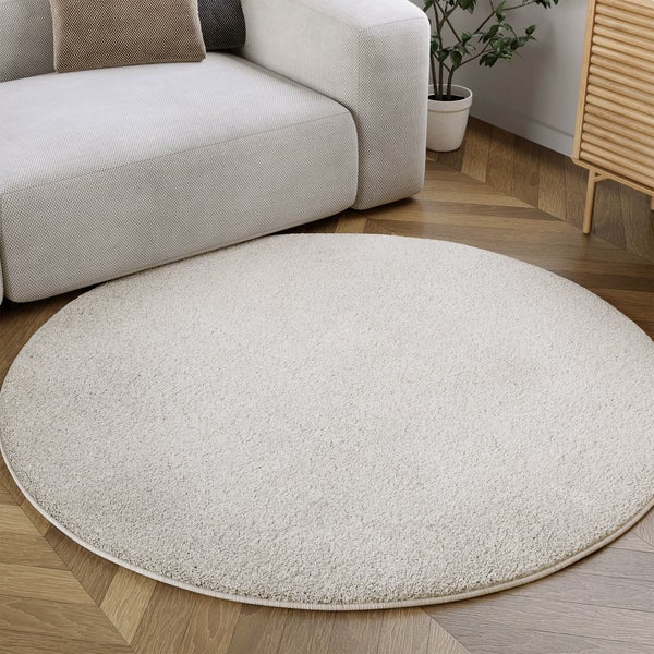 Runder, heller Hochflor-Teppich auf Fischgrät-Parkett in einem hellen Wohnzimmer mit Sofa und Holzschrank.