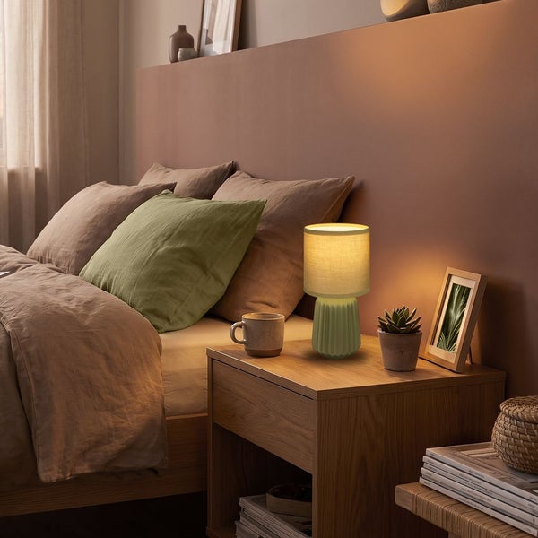 Moderne Tischlampe mit geriffeltem Sockel und Textilschirm auf einem Nachttisch in einem gemütlich eingerichteten Schlafzimmer mit Bett.