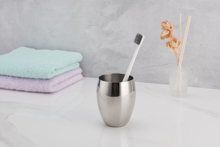 Zahnputzbecher aus Edelstahl mit Zahnbürste in modernem Badezimmer neben Handtüchern und Raumduft.