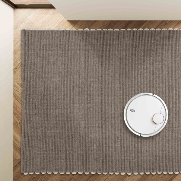 Ein weißer Saugroboter reinigt einen grauen, strukturierten Teppich auf einem Holzfußboden in Fischgrätenoptik.