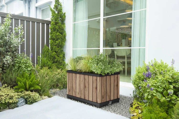 Rechteckiger Pflanzkübel in Holzoptik mit schwarzem Sockel, auf Kies in hellem Garten neben Glasfenster und grünen Pflanzen.