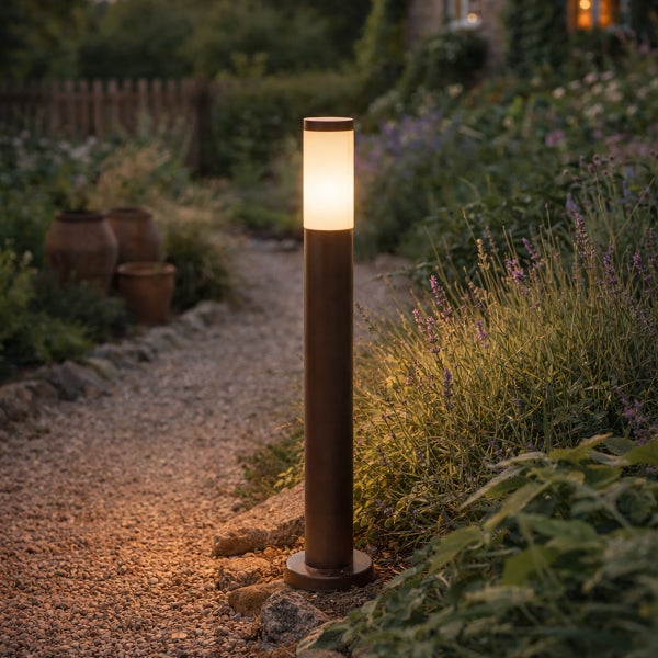 Zylindrische Pollerleuchte aus dunklem Metall beleuchtet einen Kiesweg in einem Garten mit Pflanzen und Tontöpfen.