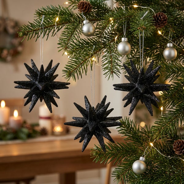 Drei hängende glitzernde Weihnachtssterne in Form von 3D Sternen als Christbaumschmuck in einem Tannenbaum mit Lichterkette.