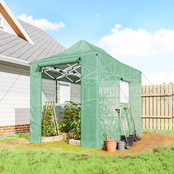Begehbares Gewächshaus mit grünem Netzbezug im Garten neben einem Haus, bepflanzt mit Tomaten, daneben stehen Gartengeräte.
