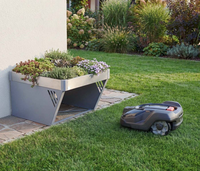Mähroboter auf grünem Rasen neben einer Metall-Garage mit bepflanztem Aufsatz in einem gepflegten Garten.