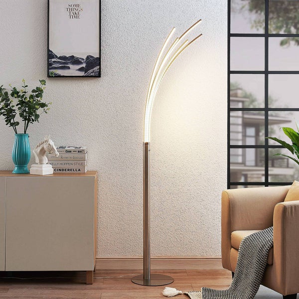 Moderne LED-Stehleuchte mit drei gebogenen Lichtelementen in einem hellen Wohnzimmer neben einem Sideboard und Sessel.