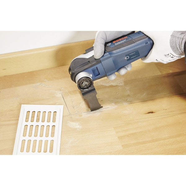 Person schneidet mit einem Multifunktionswerkzeug eine Aussparung in eine Holzoberfläche neben einem Lüftungsgitter.
