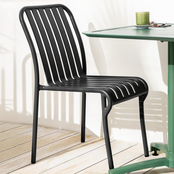 Schwarzer Gartenstuhl aus Metall mit senkrechten Latten auf einer hellen Holzterrasse neben einem grünen Tisch.