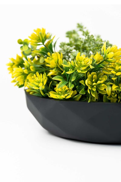 Künstliche gelbe Blumen und grünes Blattwerk in einer schwarzen, facettierten Dekoschale.