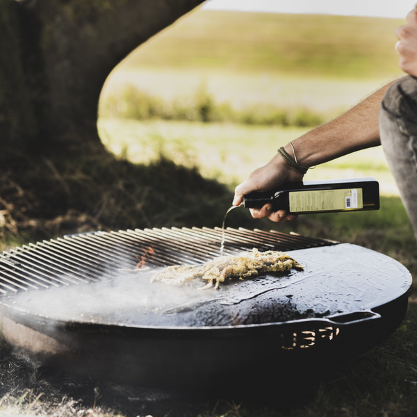 Person gießt Öl auf Grillgut auf einem Grill mit Rost und Platte im Freien.