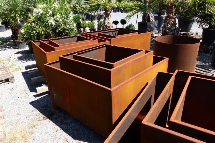 Verschiedene Pflanzkübel aus Cortenstahl in quadratischen, rechteckigen und runden Formen in einer Gartenanlage.