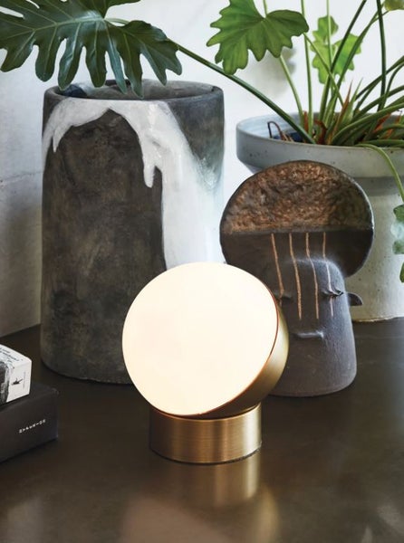 Moderne runde Tischleuchte in Metalloptik mit beleuchteter Diffusorfläche auf einer Ablage neben dekorativen Vasen und Pflanzen.