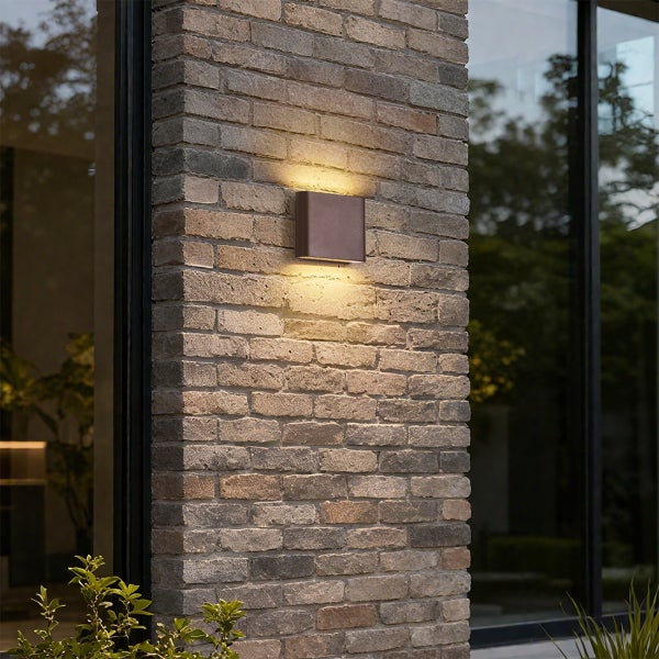 Quadratische Außenwandleuchte mit Up- und Down-Lichtaustritt an einer Backsteinwand neben Glasfenstern.