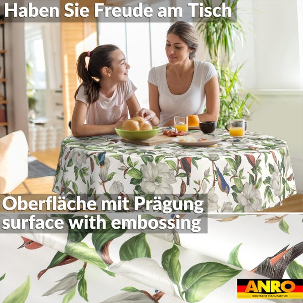 Runde Tischdecke mit Vögeln und Magnolien auf einem Esstisch in einem hellen Raum, Detail der geprägten Oberfläche und ANRO Deutsche Manufaktur Logo.