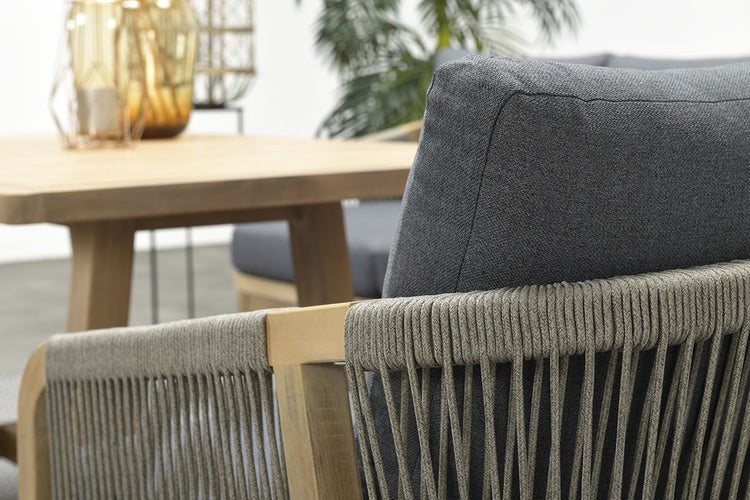 Nahaufnahme eines Gartenstuhls aus Holz mit Seilgeflecht an der Rückenlehne und grauem Polster neben einem Gartentisch aus Holz.