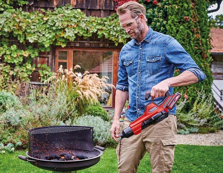Ein Mann im Garten entzündet Holzkohle in einem runden Grill mit einem roten Akku-Grillanzünder von Einhell.