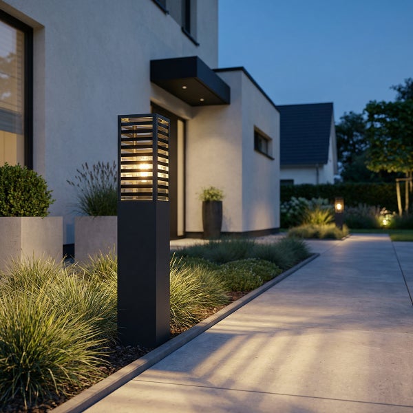 Moderne schwarze Sockelleuchte beleuchtet einen Gartenweg an einem zeitgenössischen Haus bei Dämmerung.