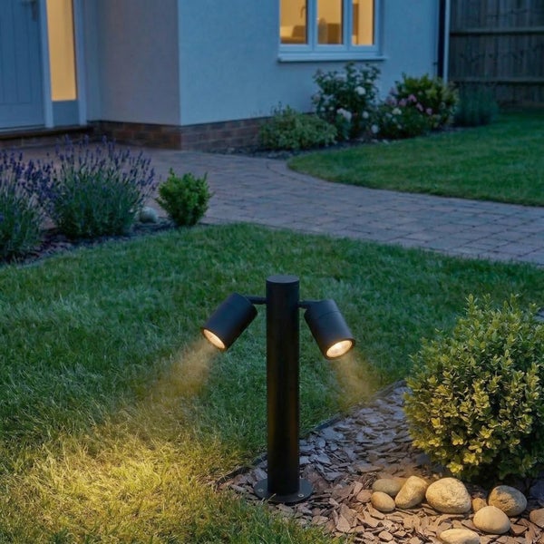 Moderne, schwarze Garten-Strahlerleuchte mit zwei Köpfen beleuchtet einen Rasen und Pflanzen in einem gepflegten Garten bei Dämmerung.