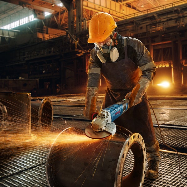 Ein Arbeiter in Schutzkleidung schleift ein Metallrohr mit einem Winkelschleifer in einer Fabrikhalle.