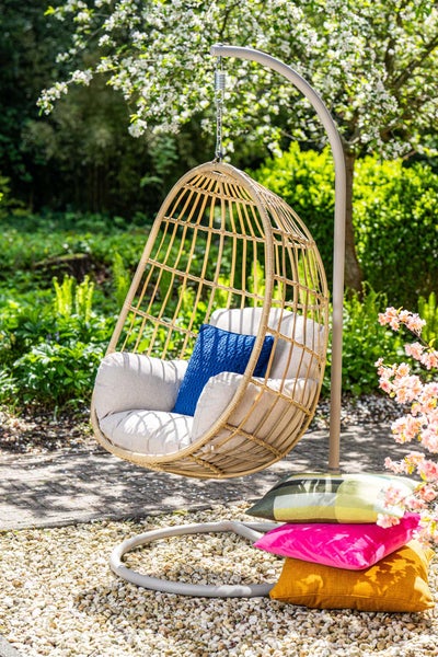 Hängesessel in Korboptik mit grauem Metallgestell und hellgrauen Sitzkissen in einem Garten mit Kiesboden.