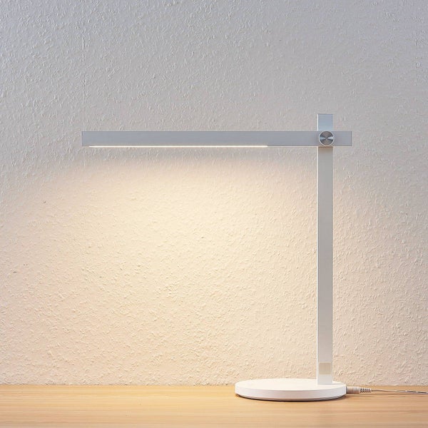 Moderne minimalistische LED Schreibtischlampe in Weiß mit schmalem Lampenarm und rundem Standfuß auf einer Holzplatte vor einer Wand.