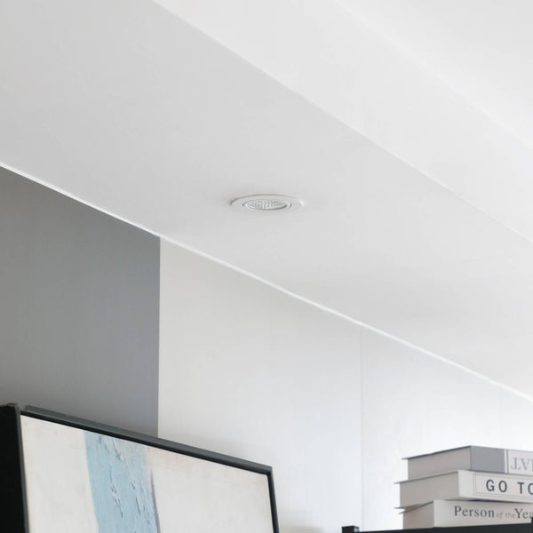 Runder LED-Einbaustrahler in einer weißen Decke über einer Zimmerecke mit Büchern und einem Bild.