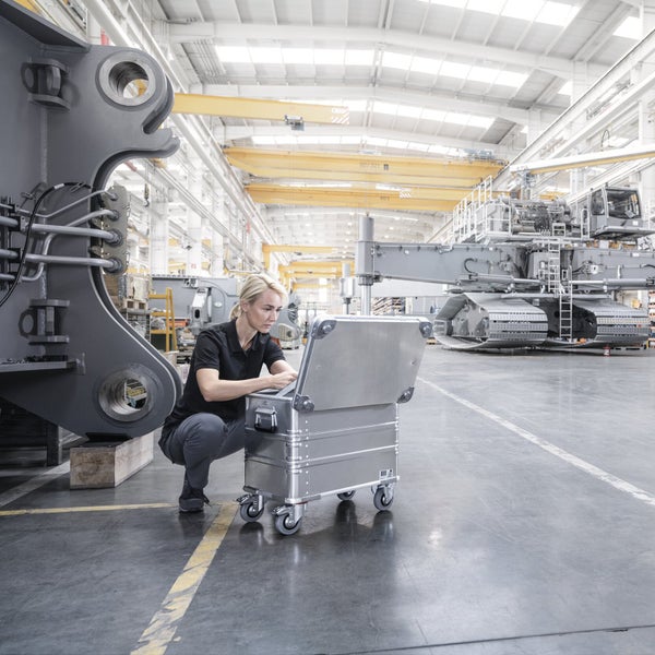 Eine Arbeiterin in einer Fabrikhalle kniet neben einer mobilen Aluminium Werkzeugkiste mit Rollen und arbeitet am Inhalt der geöffneten Box.