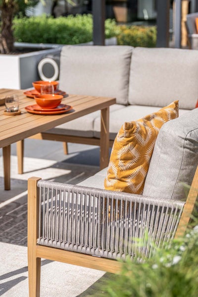 Moderne Garten-Lounge aus Holz mit hellgrauen Polstern, Seilbespannung an den Armlehnen und einem Holztisch mit orangefarbenem Geschirr.