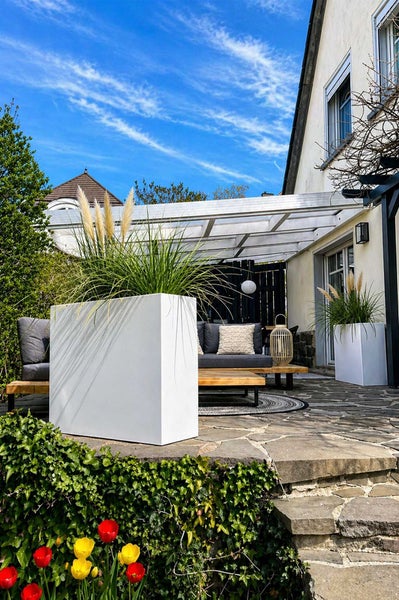 Moderne quadratische Pflanzkübel auf einer Steinterrasse mit Gartenlounge und Terrassenüberdachung.