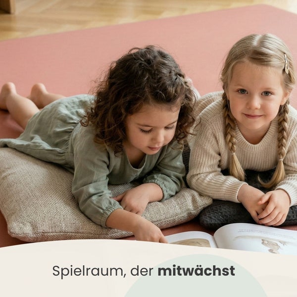 Zwei Kinder auf einer Bodenmatte und Kissen auf Holzboden in einem hellen Spielzimmer beim Buchlesen. Text Spielraum der mitwächst.