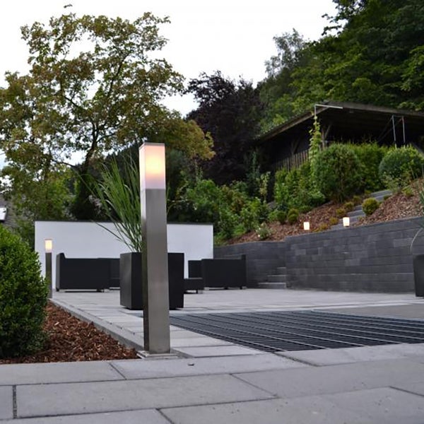 Moderne Außenleuchte aus Edelstahl auf einer gepflasterten Terrasse mit Gartenmöbeln und Steinmauer.