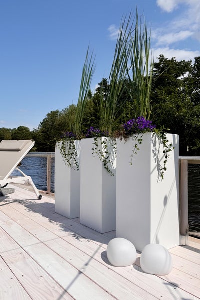 Drei hohe weiße rechteckige Pflanzgefäße auf einer hellen Holzterrasse am Wasser mit Gräsern und lila Blumen bepflanzt.