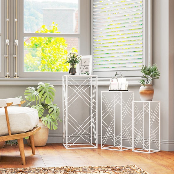 Set aus drei weißen Blumenständern aus Metall mit geometrischem Design in verschiedenen Höhen in einem hellen Wohnraum am Fenster.