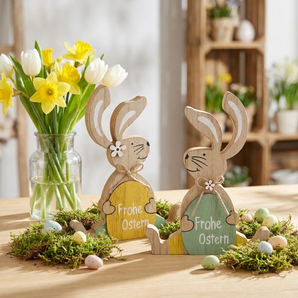 Zwei dekorative Osterhasen aus Holz mit der Aufschrift Frohe Ostern, umgeben von Moos, Ostereiern und einem Strauß Narzissen und Tulpen.