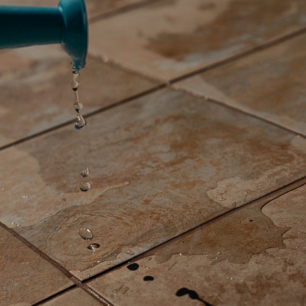 Gießkanne gießt Wasser auf einen Boden aus Steinzeugfliesen in Natursteinoptik.