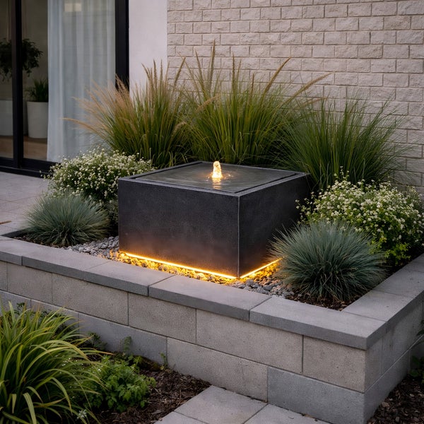 Moderner quadratischer Gartenbrunnen in Dunkelgrau mit LED-Sockelbeleuchtung, eingebettet in ein Beet mit Ziergräsern und Pflanzen.