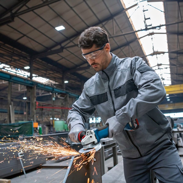 Handwerker mit Schutzausrüstung nutzt einen Akku-Winkelschleifer zur Metallbearbeitung in einer Industriehalle mit Funkenflug.