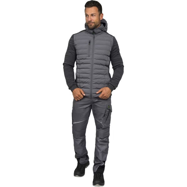 Mann in grauer Arbeitskleidung bestehend aus einer Hybridjacke mit Kapuze, einer Arbeitshose mit Taschen und schwarzen Arbeitsschuhen.
