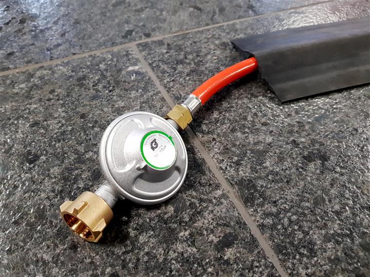Gasdruckregler 50 Millibar mit Gasschlauch und Messinganschluss auf einem Steinboden.