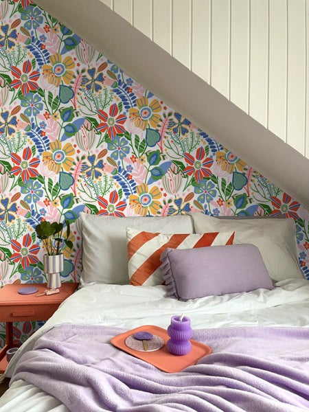Helles Schlafzimmer mit farbenfroher Blumentapete. Ein Bett mit weißem Bettzeug und lila Decke steht unter einer weißen Dachschräge.