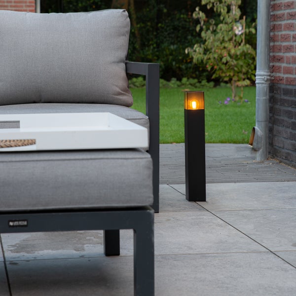 Moderne schwarze Sockelleuchte auf einer Steinterrasse im Garten neben grauen Loungemöbeln.