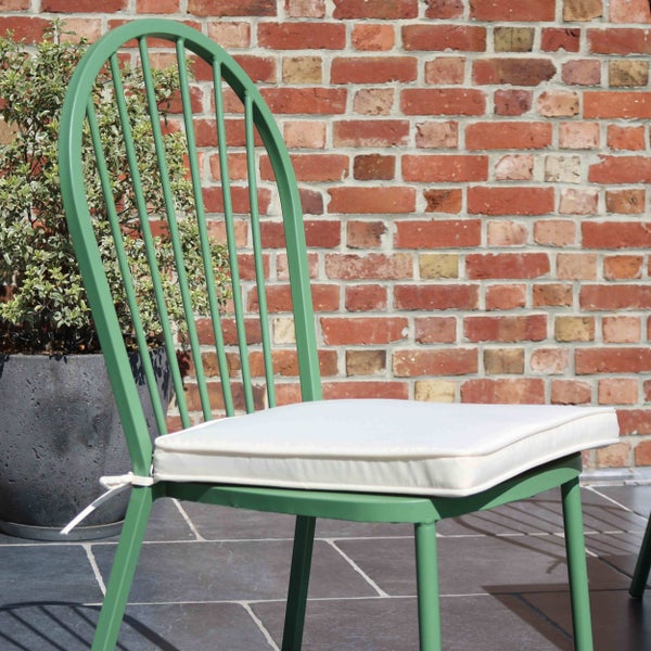 Grüner Gartenstuhl aus Metall mit weißem Sitzkissen auf einer Terrasse vor einer Backsteinwand.
