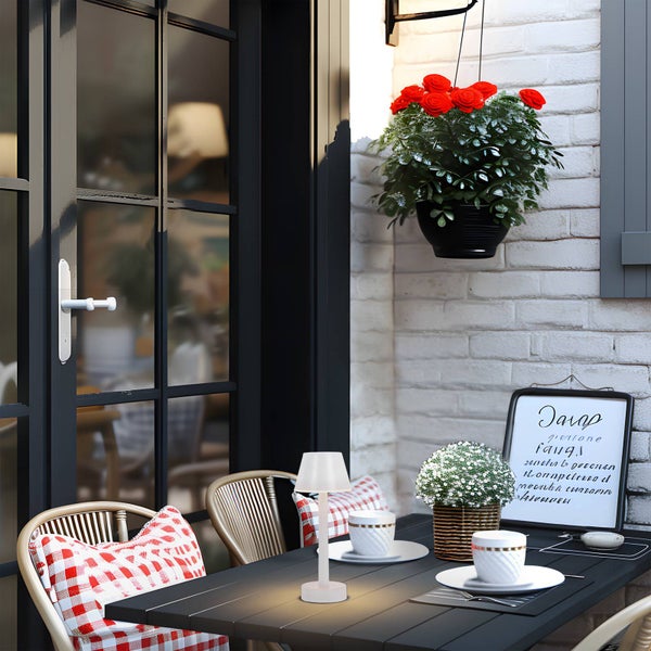 Kleine, weiße, kabellose LED-Tischleuchte auf einem dunklen Gartentisch in einer gemütlichen Außenszene mit Stühlen und Blumen.